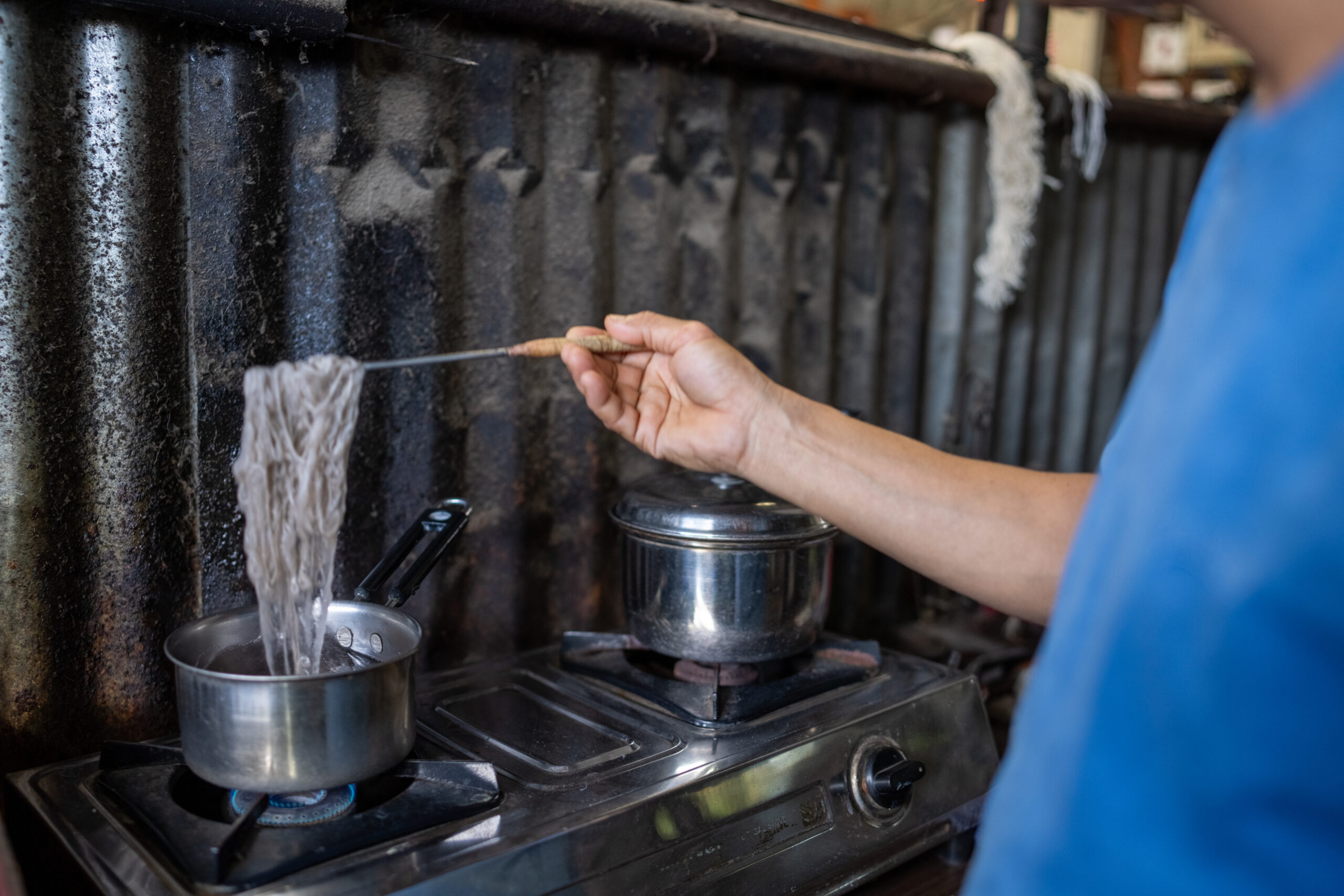 A small sample of yarn being dyed in grey colour using traditional techniques, submerged in a steaming dye bath. A wooden-handled tool lifts the freshly dyed fibers, highlighting their texture transformation in an artisanal workshop."
