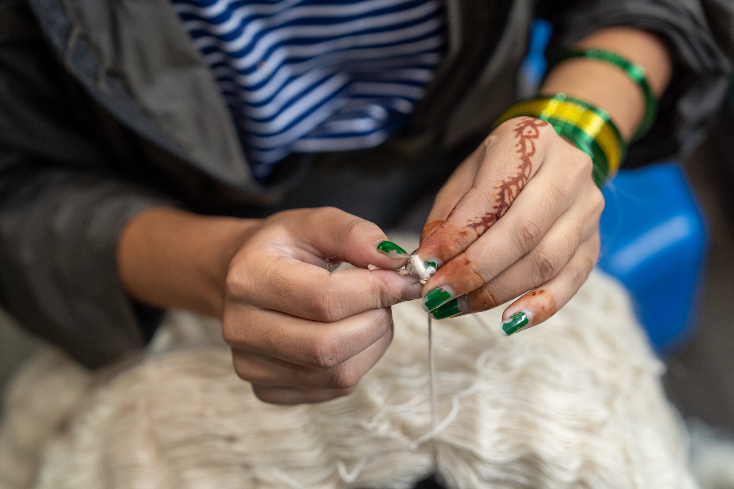 Artisan hand-knotting wool yarn for a luxury rug—close-up of skilled craftsmanship with henna-decorated hands, green bangles, and hand-dyed fibres in a traditional rug-making workshop.