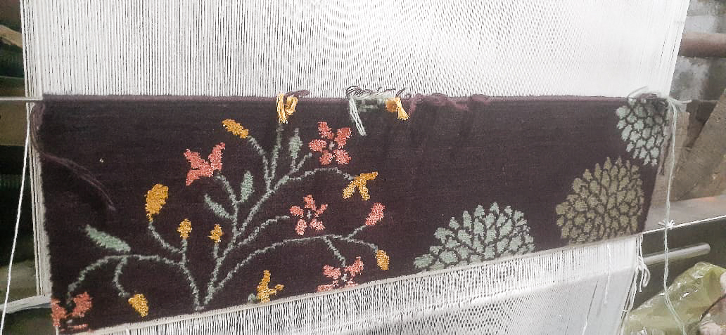 Hand-knotted floral rug in progress on loom, featuring pastel pink, yellow, and green botanical motifs on a deep plum background.