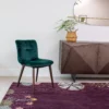 Contemporary interior with a plum floral-patterned rug, dark green velvet chair, geometric wooden sideboard, and oval brass-framed mirror—styled with a wicker vase and fern against a white wall.