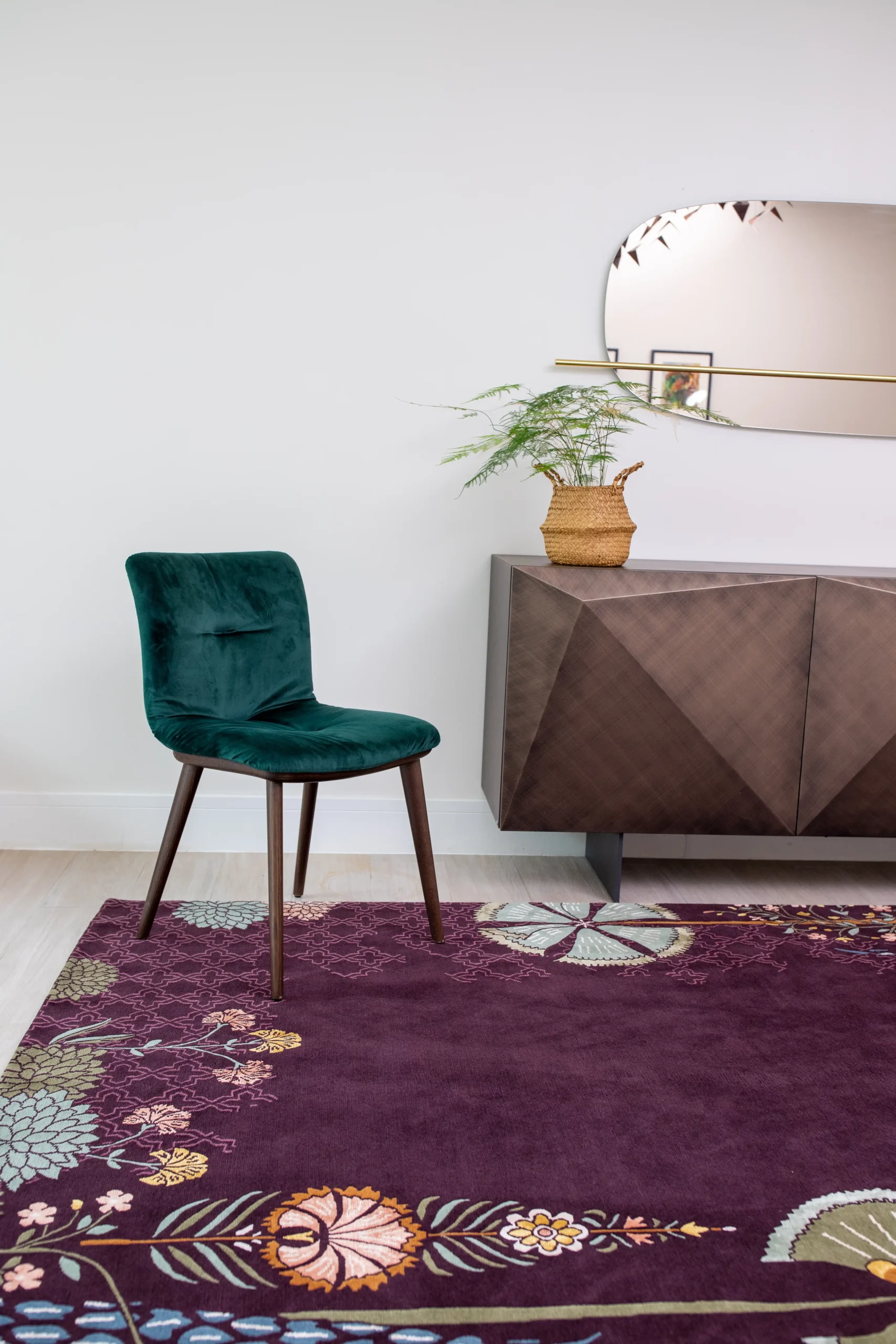 Contemporary interior with a plum floral-patterned rug, dark green velvet chair, geometric wooden sideboard, and oval brass-framed mirror—styled with a wicker vase and fern against a white wall.