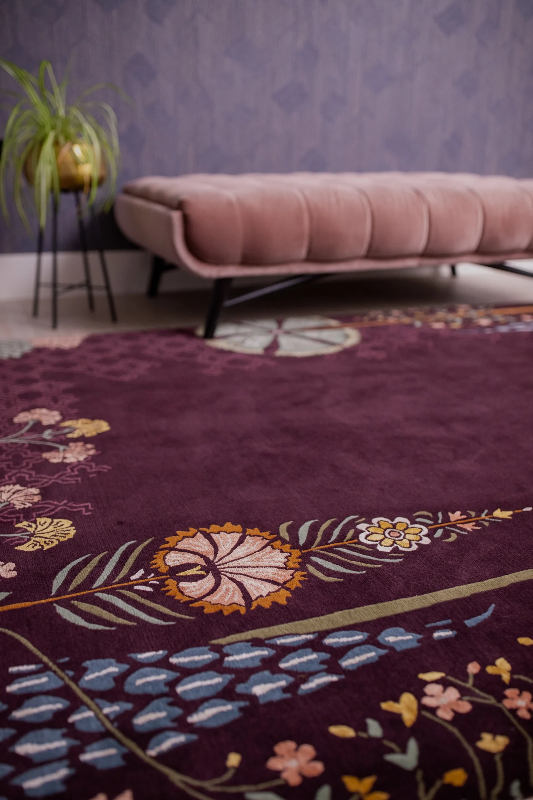 ​Plum floral-patterned rug styled with a blush velvet bench and gold planter against a soft purple textured wall— luxury interior setting with botanical detailing.
