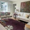 Bright, contemporary living room with a plum floral-patterned rug, white sofa, patterned accent chairs, and a modern ottoman—styled with fresh flowers, large artwork, and ample natural light from French doors.