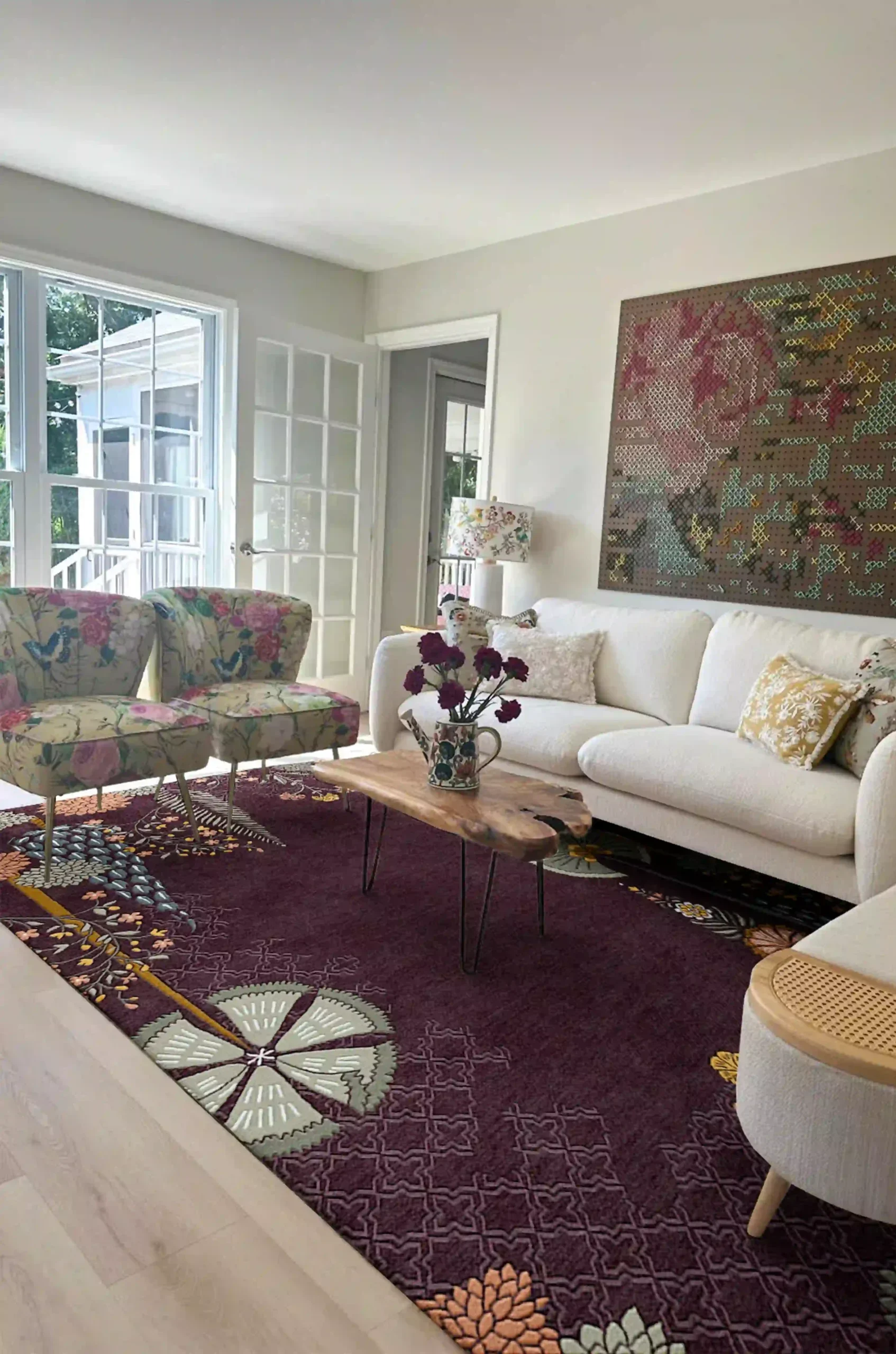 Bright, contemporary living room with a plum floral-patterned rug, white sofa, patterned accent chairs, and a modern ottoman—styled with fresh flowers, large artwork, and ample natural light from French doors.