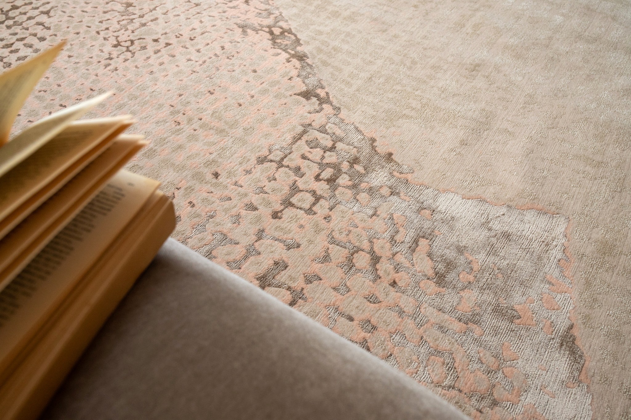 Close-up of a softly textured neutral rug in warm beige and muted blush tones, styled with a stack of art and design books and soft natural light, highlighting intricate surface details and understated elegance.